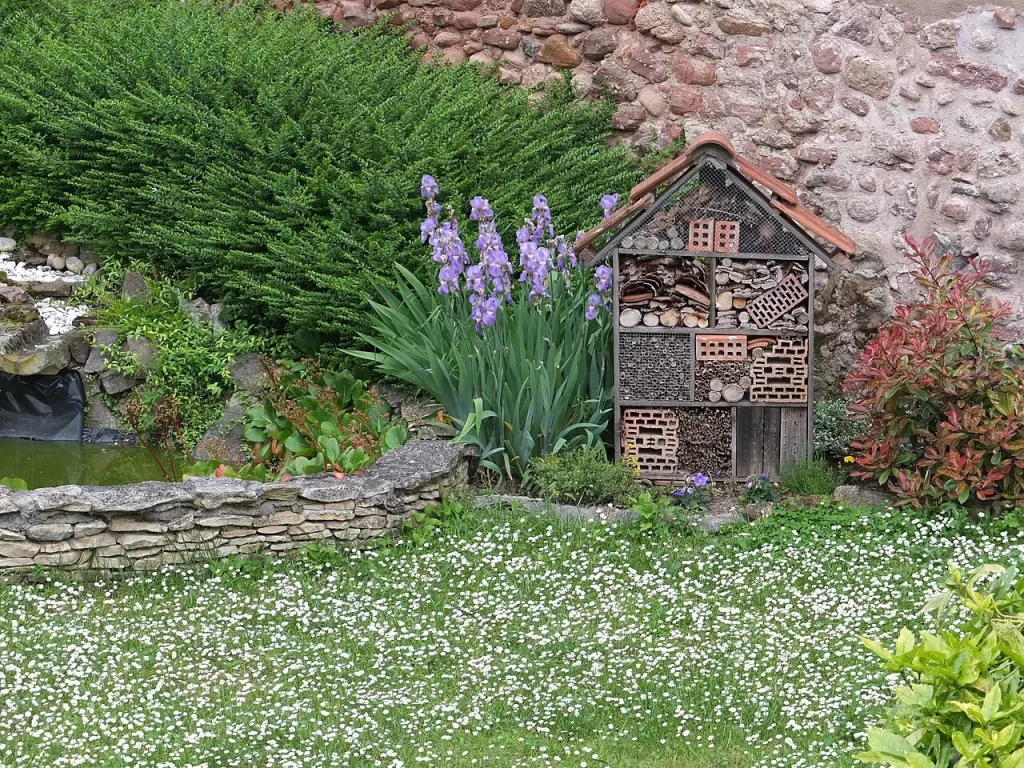 Hôtel à insectes en bois à toit de tuiles, rempli de briques, rondins et pommes de pin, dans un jardin fleuri de Guémar en Alsace
